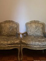 Front view of a pair of vintage armchairs with carved wooden frames and cream floral upholstery, placed side by side against white wall.