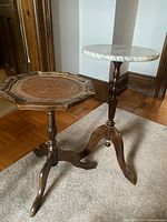 Two vintage side tables positioned on carpet and parquet floor; one with a round white marble top and the other wooden with carved tray top.