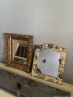 Two vintage mirrors on a surface, one ornate gold frame and one painted scenic frame.