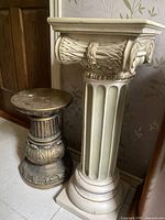 Photo showing pair of vintage plaster pedestals side by side against wallpapered wall, taller pedestal in ivory with classical fluted design and shorter darker decorative pedestal with carved detail