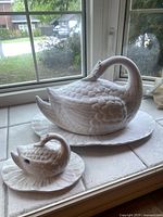 Photo showing the pair of white ceramic swan-shaped tureen and gravy boat with plates set on a tiled windowsill.