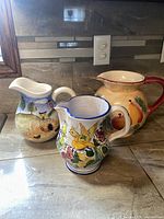 All three ceramic pitchers displayed together on a kitchen counter showing varied hand-painted farm, floral, and fruit designs.