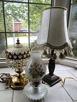 Set of 3 vintage lamps displayed on a tiled windowsill. Included is a tall lamp with a tassel-trimmed cream fabric shade, a milk glass lamp with floral hand-painted hurricane shade, and a brass lamp with crystal hanging accents.