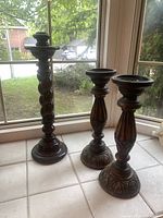 Photo showing three dark wood candle holders placed on a windowsill, one taller with barley twist design and two shorter with carved geometric details and flared carved bases.