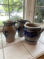 Set of three pottery pieces on tiled surface by window with natural light, showing pot with two handles and two pitchers.