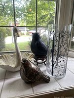 Four bird-themed decor items arranged on a windowsill including a glass vase with black tree and bird design, a carved wooden swan, metallic bird figurine, and dark bird on wood stand.
