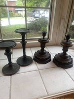 Four dark candle holders displayed on a white tile windowsill. Two are a matching pair with broad, ornate bases and thicker stems. Two are a pair with hammered texture and slimmer, taller stems, one taller than the other.