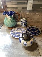 Five-piece ceramic set on counter: green and blue ceramic pitcher, two blue and white plates, small blue and white sugar bowl with lid and spoon, and lidded container.