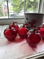 View of 8 red hollow glass apples with stems and hooks on a white surface beside a ceramic planter showing floral design.