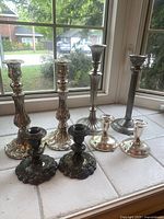 Front and side views of all eight candle holders arranged on a tile surface by a window. Three matching pairs and two individual pieces vary in height and metal finish.