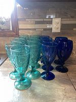 Photo showing 9 teal pressed glass goblets with embossed fruit pattern and 6 cobalt blue wine glasses, arranged on a countertop, highlighting the colors and shapes.