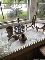 Photo showing items on window sill including pair of brass display easels, silver-tone goose figure, two brass pots, a glass candle plate, and small bag of crystals.