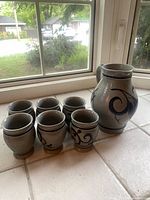 Photo showing the full set of one large Gerzit pottery pitcher and six matching glasses with blue swirl design on grey crackled finish.