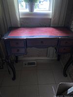 Front view of the vintage wooden desk showing red top surface with black border and five drawers in front with carved cabriole legs.