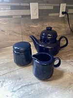 Three-piece tea set including teapot, sugar container, and cream pitcher, all in glossy cobalt blue ceramic displayed on kitchen countertop.