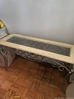 Full view of the narrow hallway table showing the cream-colored stone top with an inset glass panel and decorative metal scrolling base against a light wall and parquet floor.