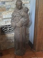Full view of vintage concrete statue of Saint Antony holding baby Jesus, showing entire figure and base, placed near stone wall and wood paneling.