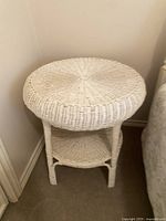 Full front view of vintage white wicker side table in corner of room showing circular top surface and lower shelf with four legs.