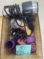 BaByliss hair dryer with wrapped cord and attached plug, placed inside a container along with multiple colorful hair curlers, electric adapters, and a large black diffuser attachment.