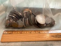 Full view of ziplock bag with multiple assorted foreign coins resting on a table with a ruler for size reference.
