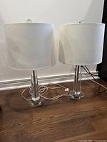 Two table lamps standing on wooden floor against white wall, showing their beige fabric shades and clear acrylic bases with cords and switches visible.