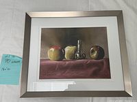 Framed still life print showing three apples and a central metallic object on a textured cloth, captured with a teal note indicating 'still life 70 artwork 18 x 21'.