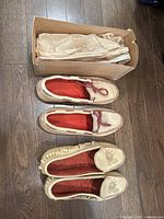 Two pairs of moccasin slippers displayed beside original box with tissue paper inside, showing cream leather with red linings and details.