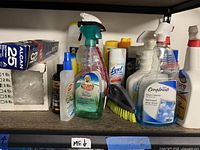 Shelf with multiple cleaning product bottles including Mr. Clean, Lysol, Compliments, and spray bottles along with cleaning brush and aluminum foil box.