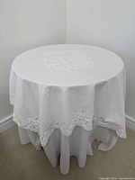 Full view of round side table covered with white embroidered cloth, showing draping fabric to floor