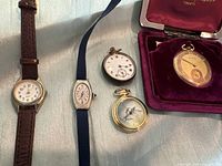 Five watches laid out on fabric: two wrist and three pocket watches, one in red velvet box