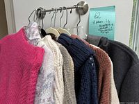 Over 10 pieces of mixed knit sweaters in different colors hanging on a rod including bright pink, cream with pattern, beige, navy blue, rust brown, and grey.