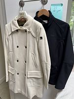 Cream double-breasted coat and black Tilley jacket hanging side-by-side on hooks