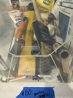 Mixed painting and repair tools including paint applicators, brushes, plastic drop sheet, filling compound and scrapers in a clear container.