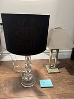 Two modern table lamps on the floor, one with clear glass base and black shade, the other with beige metal base and beige shade.
