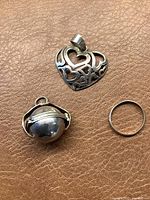 Two silver pendants and a ring laid on leather surface. Heart-shaped pendant features 'LOVE' cut-out design. Spherical locket pendant has hinged clasp.
