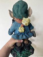 Back view of resin garden gnome showing green hat with yellow pom-poms, blue outfit, and black boots standing on grass-like base. Broken and worn.