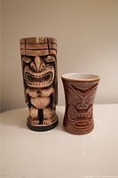 Two tiki style glasses side by side, one beige carved tiki figure glass and one brown/red ceramic tiki face glass.