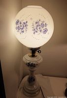 Vintage milk glass globe lamp illuminated in a corner, showing the blue floral designs on the globe and the base of the lamp.