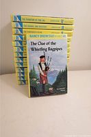 Photo showing a stack of 13 yellow-spined Nancy Drew mystery books with 'The Clue of the Whistling Bagpipes' book held in front.