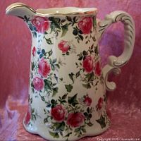 Front angled view of vintage porcelain flower jug showing rose pattern, ornate handle and gold trim.