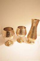 Three brass items: one vase and two pedestal glasses with engraved patterns on a white surface under indoor lighting.