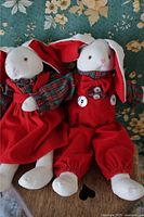 Front view of two plush bunnies dressed in red velvet Christmas-themed outfits with plaid details, seated on floral fabric.