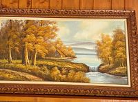 Full view of the vintage oil painting depicting an autumn landscape with water and trees in an ornate wooden frame.