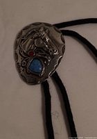 Close-up of the metal horse head medallion with turquoise and red stone inlays on black braided cord.