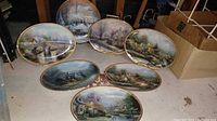 All seven Thomas Kinkade oval plates laid out on floor