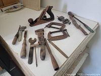 Photo showing a collection of 12 assorted antique hand tools laid out on a white surface including a wood planer, clamps, screwdrivers, pliers, files, and a scraper all with rust and wear.