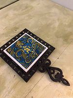 Close-up photo of a square decorative tile with elaborate blue and yellow floral patterns set into a heavy decorative metal frame with hanging hook.