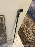 Full length photo of the hand-carved bone walking stick leaning against wall on tiled floor, showing smooth straight bone shaft with a curved and detailed handle.