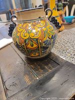 Front angle view of a round Mexican ceramic vase with vibrant hand-painted floral and geometric designs in yellow, okra, blue, and cream. Decorative black curled handles on each side.