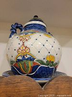 Photo of large ornamental Mexican urn showing colorful floral and geometric folk art pattern on a white ceramic base, details of lid and part of wooden shelf below.
